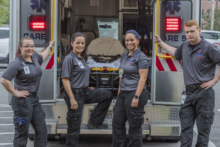 Four EMT students stand behind an Ambulance.