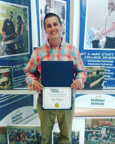 Ben Cochran is standing in front of some various college banners while holding a certificate.