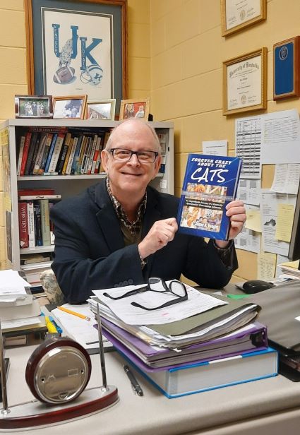 Jamie Vaught smiles for a photograph while he is sitting at his desk. He is holding up a copy of his latest book.