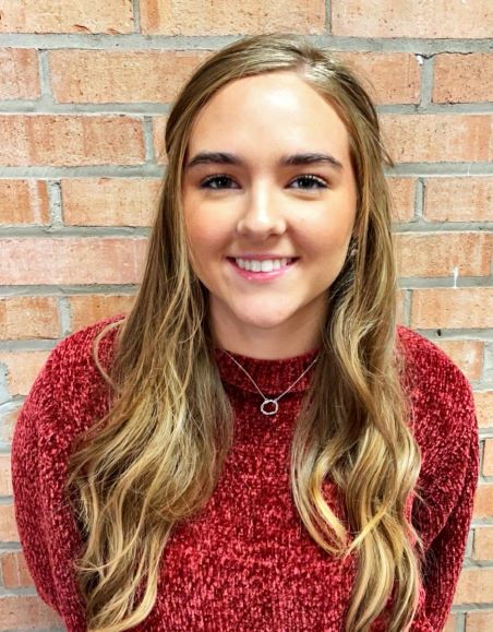 Leah Sturgill poses for a photograph with a brick wall behind her. She is wearing a red sweater.