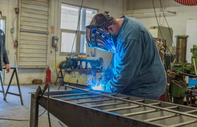 A welder is working in a shop. He is wearing a face shield and sparks are flying around his gloved hands.